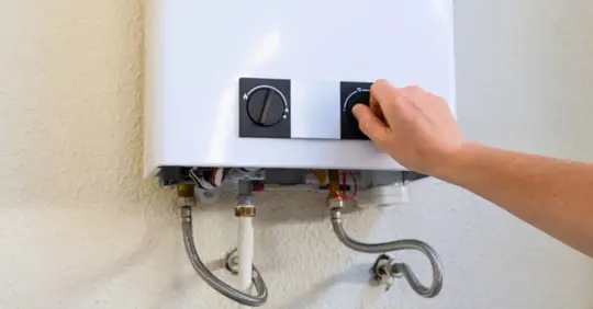 Person's hand adjusting the temperature dial on a tankless gas water heater mounted on a wall