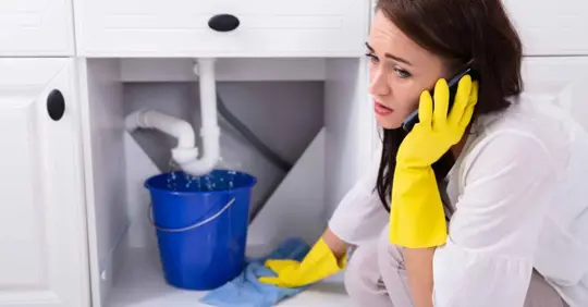 Woman Calling Plumber In Front Of Water Leaking From Pipe